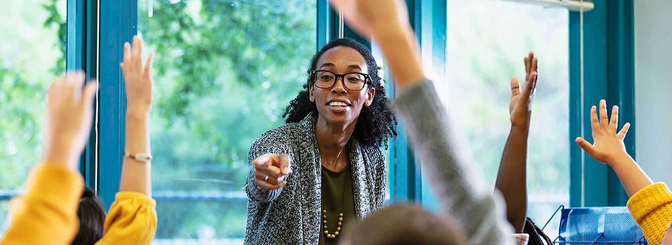 Image of a teacher pointing at students raising their hands in a clasroom